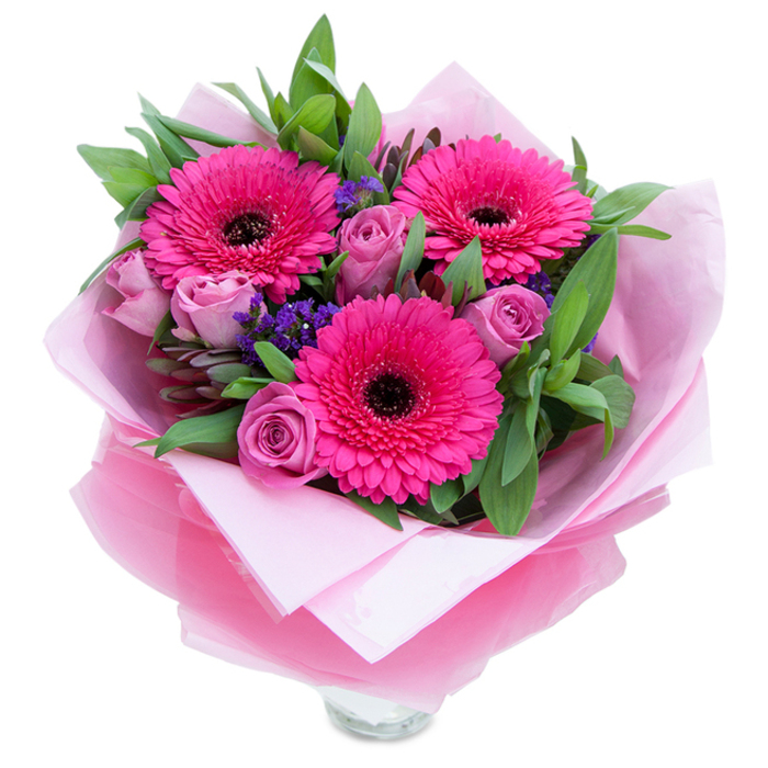 There is a charming balance of playfulness and grace in this pink-dominant bouquet, where three confident fuchsia Gerbera daisies take centre stage and command attention with their saturated colour and dark, contrasting hearts. Around them, dusty-lavender roses nestle in close, their petals forming graceful spirals that soften the arrangement and invite a tender, almost wistful response. Little clusters of purple-blue statice are dotted throughout like confetti, lending an informal, meadow-like texture that suggests a freshly gathered posy rather than something overly formal. The greenery-broad, dewy leaves suggesting salal or eucalyptus-creates a structured yet natural backdrop, and the faint aromatic notes of foliage mingle with the roses' powdery perfume. The bouquet is carefully wrapped with an inner sheet of matte pale-pink tissue and an outer layer of translucent, slightly crinkled fuchsia-pink cellophane that catches light and adds depth; the wrapping is folded with a practiced hand so the flowers feel both secure and unhurried. The top rim of a clear glass vase is visible beneath, offering a glimpse of water and promise of longevity once placed on a bedside table or hallway console. Bathed in soft, diffuse light-the kind you might notice in an afternoon delivery to Hemel Hempstead Old Town-the colours read warm and consoling. Hand-tied to feel intimate yet polished, this bouquet is ideal for intimate celebrations, a thoughtful apology, or to bring a gentle smile to a neighbour in Adeyfield, reflecting the kind of attentive craftsmanship found in local floristry.