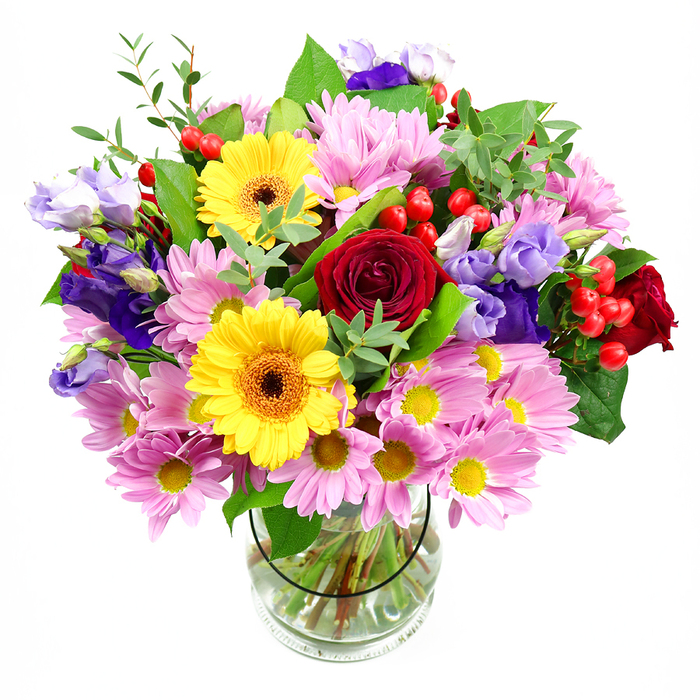 A vibrant, mixed-flower bouquet stands tall in a clear glass vase, perfectly centred against a stark white background that throws the colours into sharp relief. Deep red roses sit at the heart of the design, their velvety petals unfurling in generous layers that speak of classic romance and enduring affection. Nestled near them, two radiant yellow gerbera daisies with distinctive brown-orange centres introduce a burst of sunshine; their open, daisy-like faces bring a modern brightness that feels instantly joyful. Spilling outward, clusters of soft pink daisies or chrysanthemums form a gentle ring around the central blooms, their finely cut petals and tiny yellow centres adding delicacy, movement, and a pastel glow reminiscent of flowerbeds along the streets of Hemel Hempstead in spring. Interlaced among these are slender stems of purple and lavender lisianthus, whose bell-shaped flowers curl and twist slightly, creating shadows and depth that make the bouquet visually rich and layered. Tucked between flowers, small clusters of glossy red Hypericum berries provide unexpected texture, standing out like polished beads and adding a hint of contemporary flair. The entire arrangement is cradled in lush greenery, from large, dark leaves that give structure and weight, to lighter, more feathery greens that soften the outline and suggest a freshly picked, garden-inspired style. The clear glass vase reveals the neatly trimmed stems and sparkling water, underscoring the professional care behind the piece. The overall mood is warm, celebratory, and inviting, making this bouquet ideal for sending congratulations to a new home in Leverstock Green, marking a birthday, or simply brightening a dining table in Hemel Hempstead with a joyful, colourful presence.